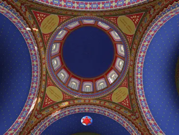 Das Bild zeigt eine Kuppel einer Synagoge von innen. Der gr&ouml;&szlig;te Teil der Decke ist blau, die verschiedenen Ornamente sind gelb und rot. 