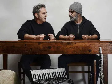 Two men are sitting next to each other at a wooden table, looking at each other, their four fists clenched on the tabletop. In front of the table are five instruments: a drum, an accordion, two flutes and a clarinet.