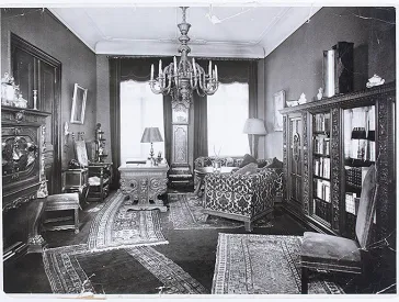 Schwarz-Weiß Bild. Ansicht eines Wohnzimmers mit schweren Holzmöbeln und mehreren Teppichen.  