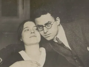 Die historische Fotografie zeigt eine junge Frau, zu deren rechter Seite ein Mann an ihren Kopf gelehnt ist. Er blickt in die Kamera und umfasst die Schultern der Frau mit seinem rechten Arm.