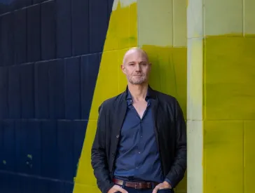 Ein Mann steht vor einer gelb-blauen Wand. Er hat seine H&auml;nde in die Hosentaschen gesteckt. 