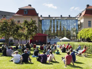 Several people are sitting on the lawn in the museum garden, looking toward the stage.