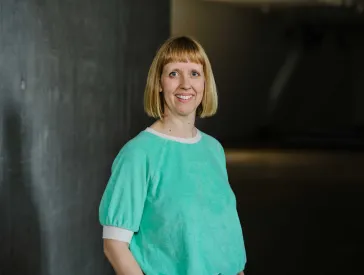 A woman with shoulder-length, blonde hair is smiling and looking directly towards the camera. Her upper body is visible. She is wearinga turquois coloured sweater.