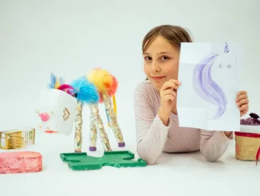 Ein Kind neben einem gebasteltem Einhorn hält eine Einhorn-Zeichnung in die Kamera