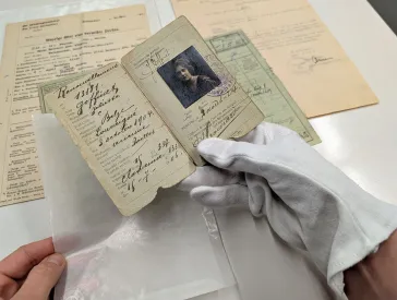 Eine Hand mit Baumwollhandschuh hält einen historischen Ausweis in die Kamera. Im Hintergrund sind weitere historische Dokumente zu sehen. 