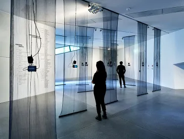 Ausstellungsansicht mit Besucher:innen, von der Decke hängen schwarze durchsichtige Stoffbahnen, daran befestigt sind Smartphones mit Klangwellen