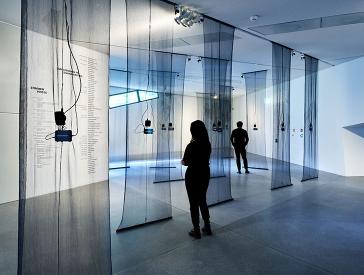 Ausstellungsansicht mit Besucher:innen, von der Decke hängen schwarze durchsichtige Stoffbahnen, daran befestigt sind Smartphones mit Klangwellen