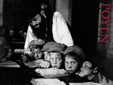 The black and white photo shows several boys in caps sitting close together at a table. In front of them are open books, behind them stands a man and points to a line of text