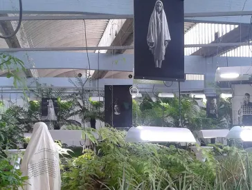 Pflanzen-Plateaus im Inneren einer großen Halle, dazwischen Schaufensterpuppen mit Modeentwürfen, darüber hängen großformatige Fotografien.