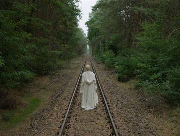 The photo shows a person from behind, dressed in a white frock. The person follows railroad tracks that run dead straight through a forest