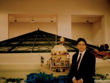 A boy in a dark suit and tie stands next to a toy ark made of wood. A rotunda is visible in the background.