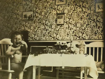  Junge sitzt nackt und etwas aufgebracht am Tisch; das Wohnzimmer ist mit einer Blumentapete tapeziert.