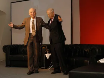 Two elderly gentlemen (Horst Eisfelder on the left, W. Michael Blumenthal on the right), both wearing suits, are standing side by side on a stage. They are facing the audience and have their arms casually around each other.