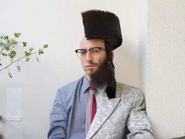 Fotografie eines Mannes, der entlang der Längsachse gleichsam in zwei Hälften geteilt ist: Auf der linken Seite trägt er Hemd und Glatze, auf der rechten Vollbart und einen (halbierten) Schtreimel auf dem Kopf