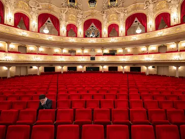 Person allein auf einem der roten Sitze im Parkett eines leeren Theaters, den Kopf in die Hände gestützt