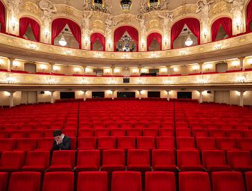 Person allein auf einem der roten Sitze im Parkett eines leeren Theaters, den Kopf in die Hände gestützt