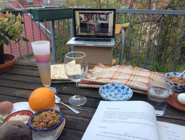 On a wooden table there are various glasses, plates with dishes, bowls and an open book. On a plate lies an orange. Behind the table there is a laptop on a cardboard box, in which a video call can be seen.