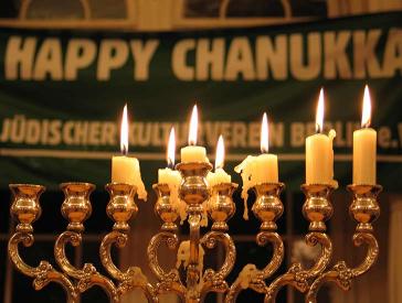 Chanukkaleuchter mit brennenden Kerzen, dahinter ein Banner mit der Aufschrift: Happy Chanukka. Jüdischer Kulturverein Berlin.