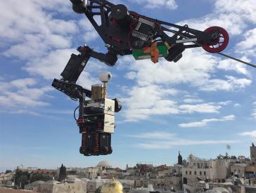 A camera mounted on a steel cable hovers before a blue sky over the rooftops of Jerusalem.