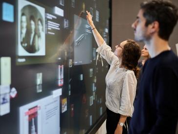 Menschen berühren eine große Touchscreen-Wand, auf der Dokumente und Objekte zu sehen sind