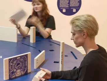 Two people are sitting at a round table in an exhibition space, looking at wooden objects.