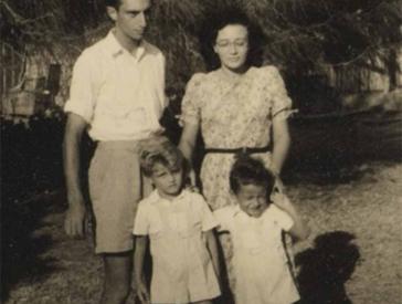 Die Aufnahme zeigt einen Mann, eine Frau, vor ihnen zwei Kinder, alle stehen im Freien. Die beiden Jungs und der Mann tragen kurze Hosen, die Frau ein kurzärmeliges Kleid mit floralem Muster.