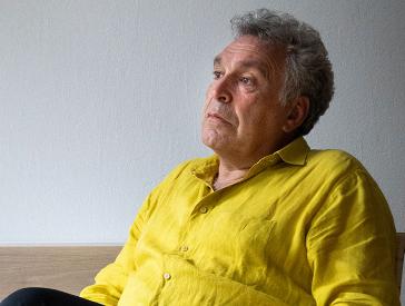 Photo of Frédéric Brenner in half profile, sitting, wearing a yellow shirt