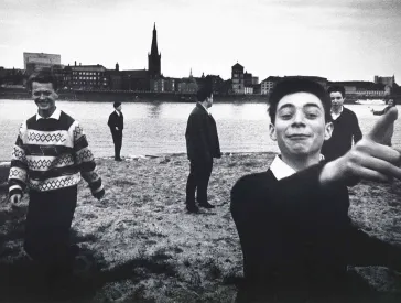 Black and white photograph: Six young people stand on the banks of a wide river. They are not standing together, but spread out across the entire picture. Some are looking towards the viewer, others across to the city that stretches along the opposite side of the river. The group makes an exuberant impression.