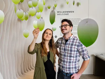 Ein Mann und eine Frau schauen sich grüne Papierblätter an, die an Fäden von oben runterhängen.