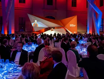 Festsaal mit Gästen an Tischen und einer ausgeleuchteten Bühne im Hintergrund.