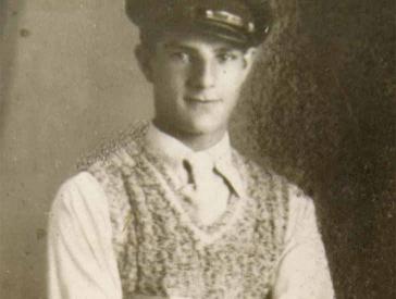 Portrait of a standing young man in a sailor's cap and a hand-knit sweater. He has a friendly look on his face and a delicate build.