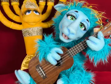 A blue doll with big eyes plays guitar, behind her a doll in the shape of a menorah.