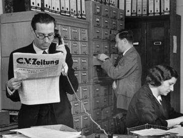 A man in a suit is reading the C.V. newspaper while standing and talking on the phone, in the background a woman at a desk and a man at a filing cabinet.