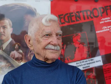 Portrait of an elderly man (Harry Raymon) with gray hair and mustache, a film poster can be seen in the background.
