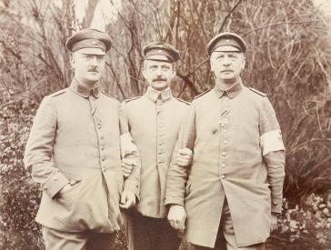 Black and white photograph with three uniformed soldiers standing in front of a green area.