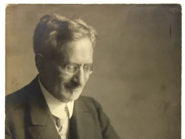 Schwarz-Weiß-Fotografie eines Mannes im Anzug und mit Brille, zeitungslesend an einem Tisch sitzend.