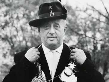 Schwarzweiß-Fotografie: Ein Herr mit Jagdhut und Anzug präsentiert eine Kette mit zahlreichen Anhängern.