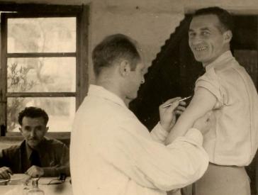 Black-and-white photograph of a man holding a scalpel to the upper arm of another man who is smiling for the camera