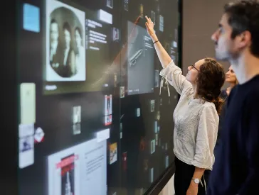 Medienstation mit einem gro&szlig;en Bildschirm vor der drei Personen stehen