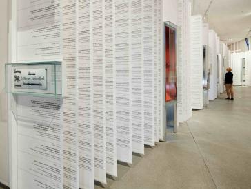 Long white paper banners with text hang from the ceiling interrupted by showcases, in the background stands a blonde woman