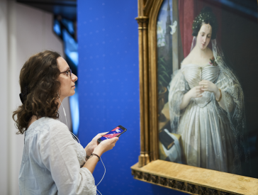 Young woman stands with smartphone and headphones in front of portrait of Albertine Heine as bride of August Theodor Kaselowsky.