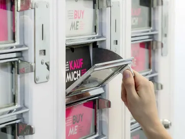 Eine Hand öffnet eines der Fächer des "Kunstautomaten", der Text in dem Fach lautet "Kauf mich".