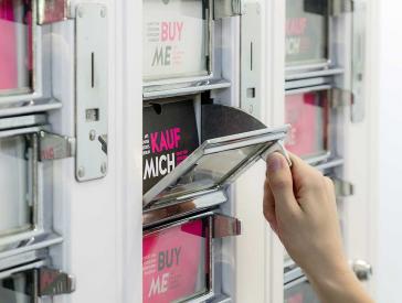 Eine Hand öffnet eines der Fächer des "Kunstautomaten", der Text in dem Fach lautet "Kauf mich".