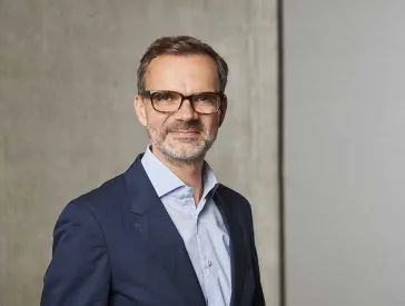 Ein Mann mit braunen Haaren und Brille steht vor einem Betonhintergrund. 