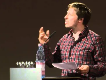 Ein Mann auf einer B&uuml;hne hinter einem Pult h&auml;lt eine Rede.