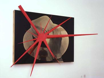 The large-scale artwork shows a highly magnified photo of a hat and a hand about to lift the hat. On the photo is mounted a red three-dimensional virus-like object