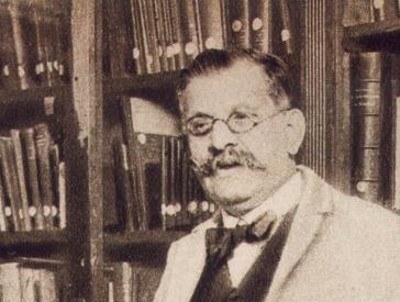 Schwarz-weiß-Foto: Magnus Hirschfeld mit Brille, Schnurbart und weißem Kittel. Er steht vor einem Buchregal und schaut freundlich in die Kamera.