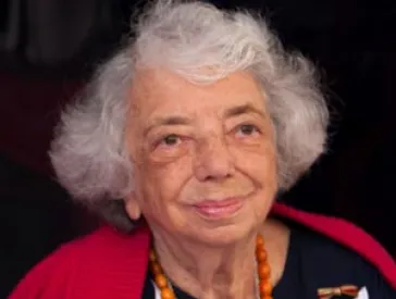 Woman (Margot Friedländer) with white hair and striking amber necklace looks friendly into the camera.
