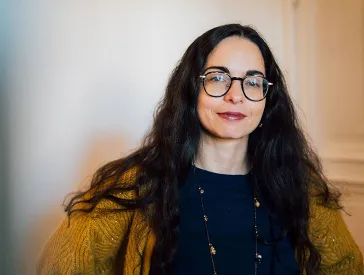 Eine Frau mit dunklen, langen Haaren und Brille schaut freundlich in die Kamera.