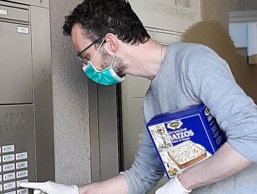 A man wearing a surgical face mask with a package of matzo under his arm rings a doorbell.